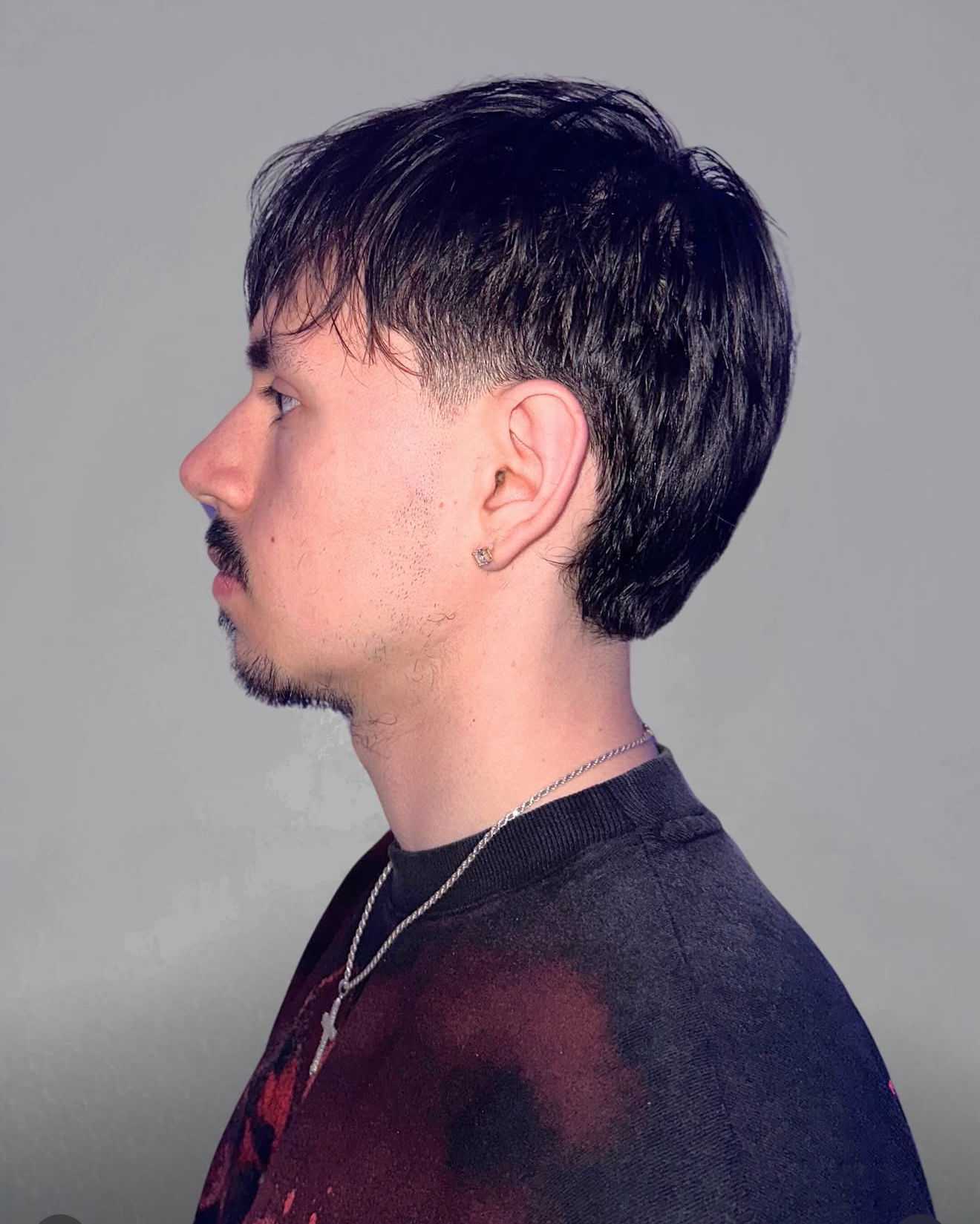 Man with short hair and necklace, side profile against gray background.