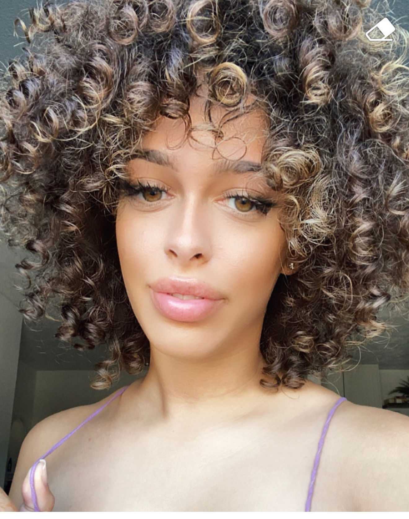 Curly-haired person smiling, wearing a purple top, with a neutral background.