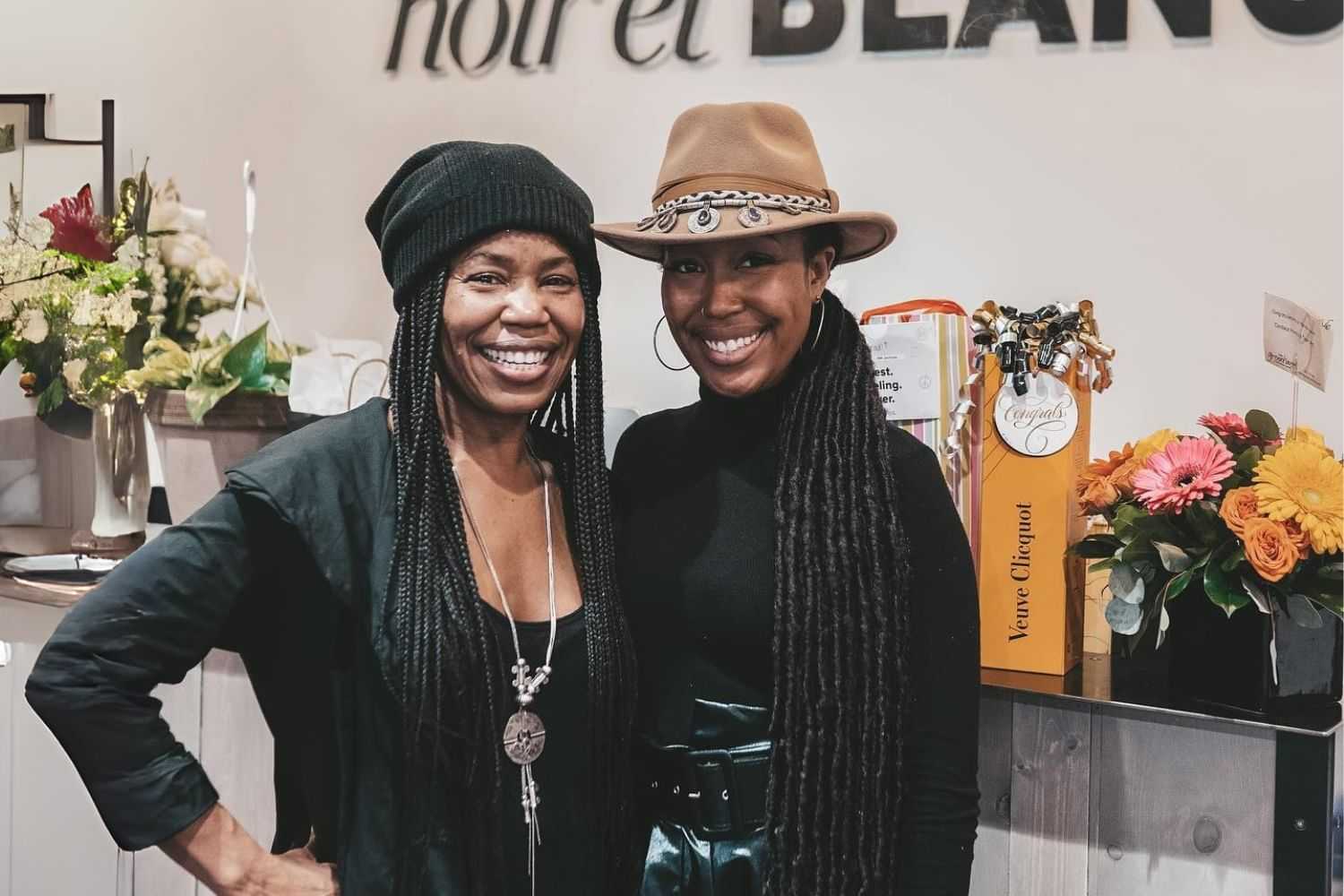 Two women smiling, standing by flowers and a gift in a stylish interior.